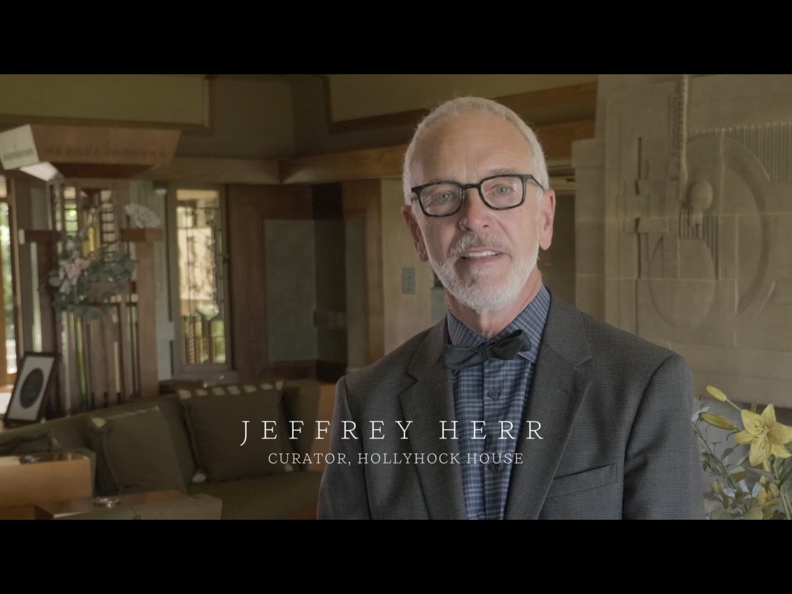 Jeffrey Herr, curator of the Hollyhock House is wearing a suit, bowtie and black eyeglasses and staring directly at the camera.  He is standing in a large room with sand colored walls and a green couch behind him.